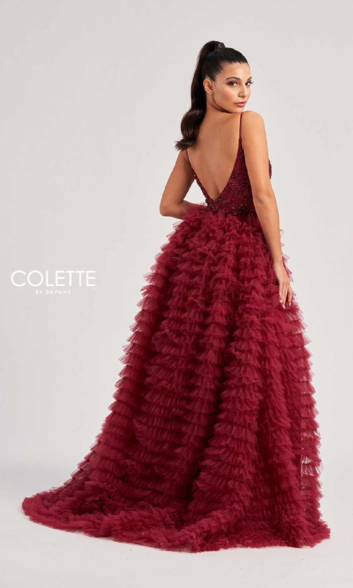 A woman wearing the Colette CL8330 long prom dress—a burgundy gown with a tiered ruffled skirt and deep V-back—stands looking over her shoulder against a plain background.